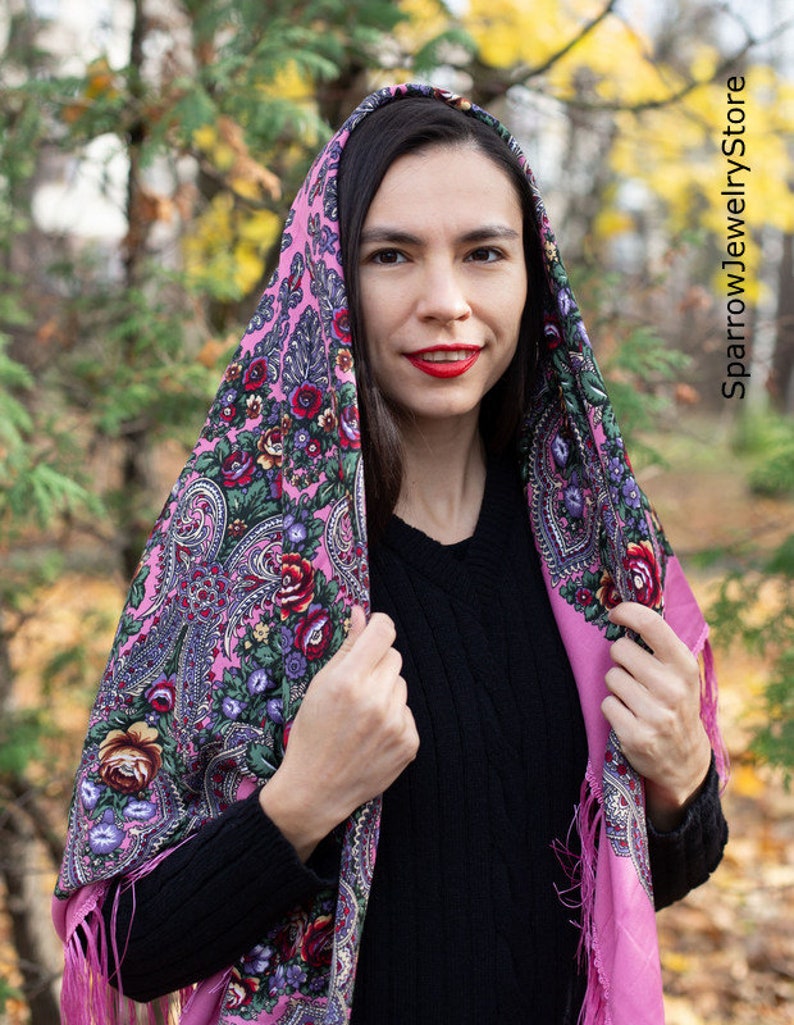 May include: A pink patterned headscarf with floral designs and fringe. The scarf is worn by a person with long dark hair and red lipstick.