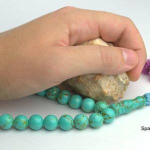 May include: A turquoise blue prayer bead mala with a light blue tassel. The mala is made of round beads with a marbled pattern. The mala is lying on a white surface with a seashell and purple flowers.