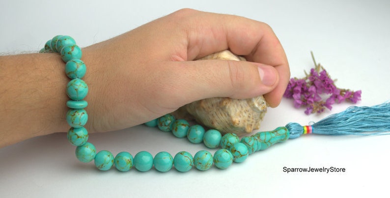 May include: A turquoise blue prayer bead mala with a light blue tassel. The mala is made of round beads with a marbled pattern. The mala is lying on a white surface with a seashell and purple flowers.