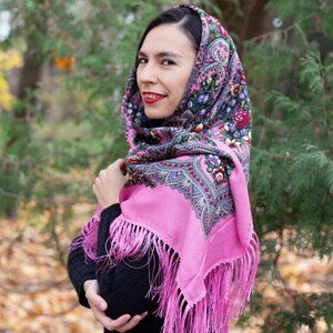 May include: A pink headscarf with a floral pattern and fringe. The scarf is worn by a person with dark hair and red lipstick. The background is a blurred image of trees and leaves.