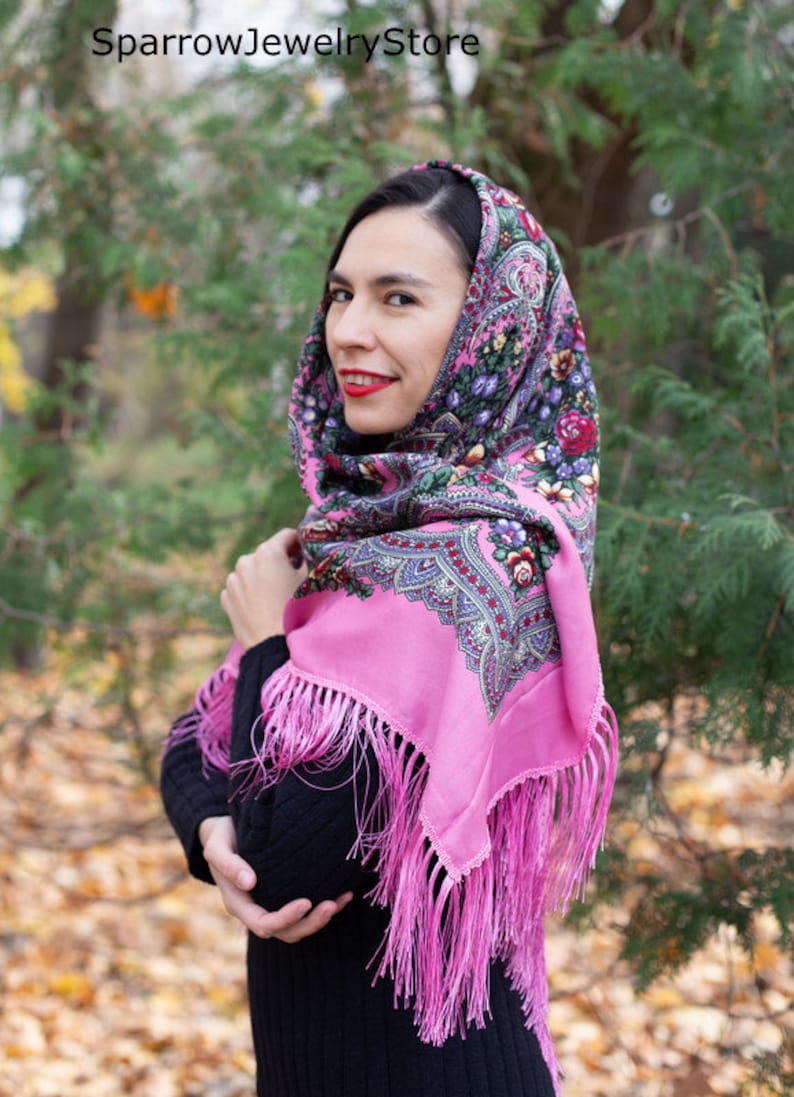 May include: A pink headscarf with a floral pattern and fringe. The scarf is worn by a person with dark hair and red lipstick. The background is a blurred image of trees and leaves.