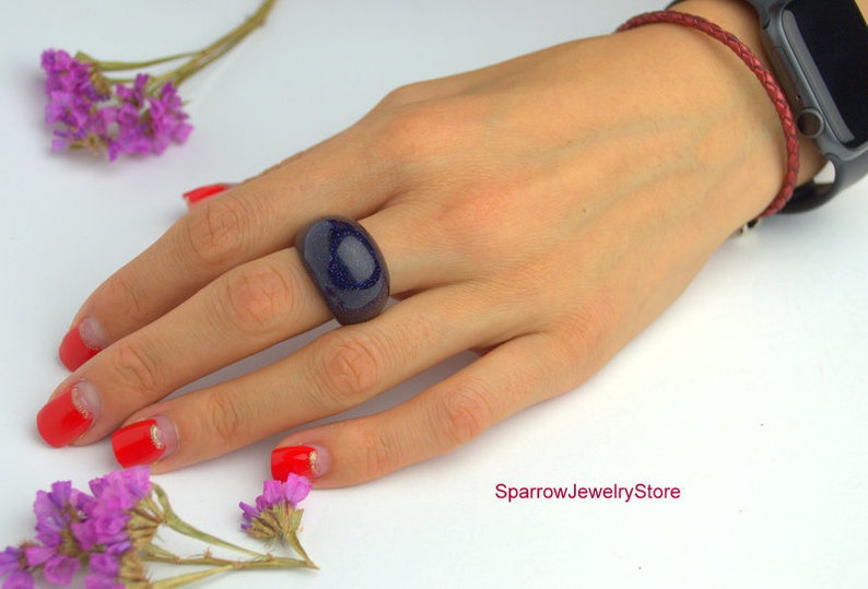 May include: A close-up of a hand wearing a dark blue ring with a sparkly finish. The ring is on the middle finger of the hand. The hand is wearing a red braided bracelet on the wrist.