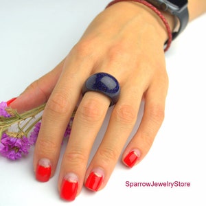 May include: A dark blue ring with a speckled texture. The ring is made of resin and is worn on a finger with red nail polish.