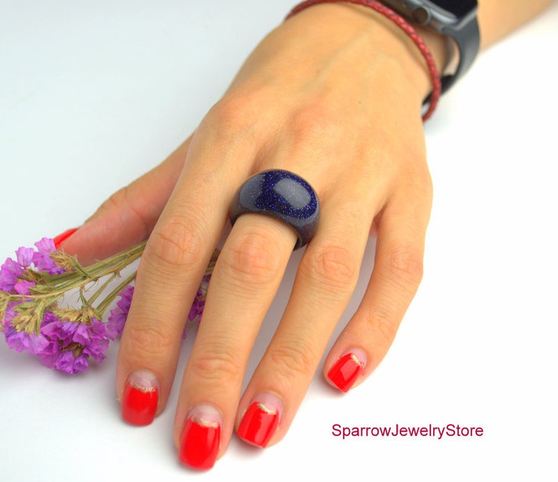May include: A dark blue ring with a speckled texture. The ring is made of resin and is worn on a finger with red nail polish.