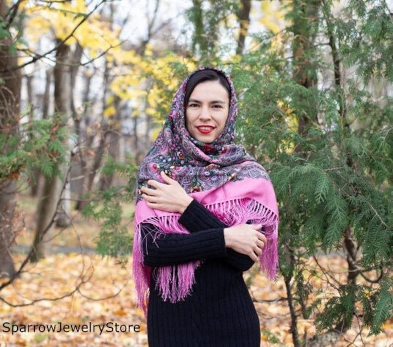May include: A woman wearing a pink and black patterned headscarf with fringe. The scarf is wrapped around her head and neck. She is standing in a forest with fall foliage.