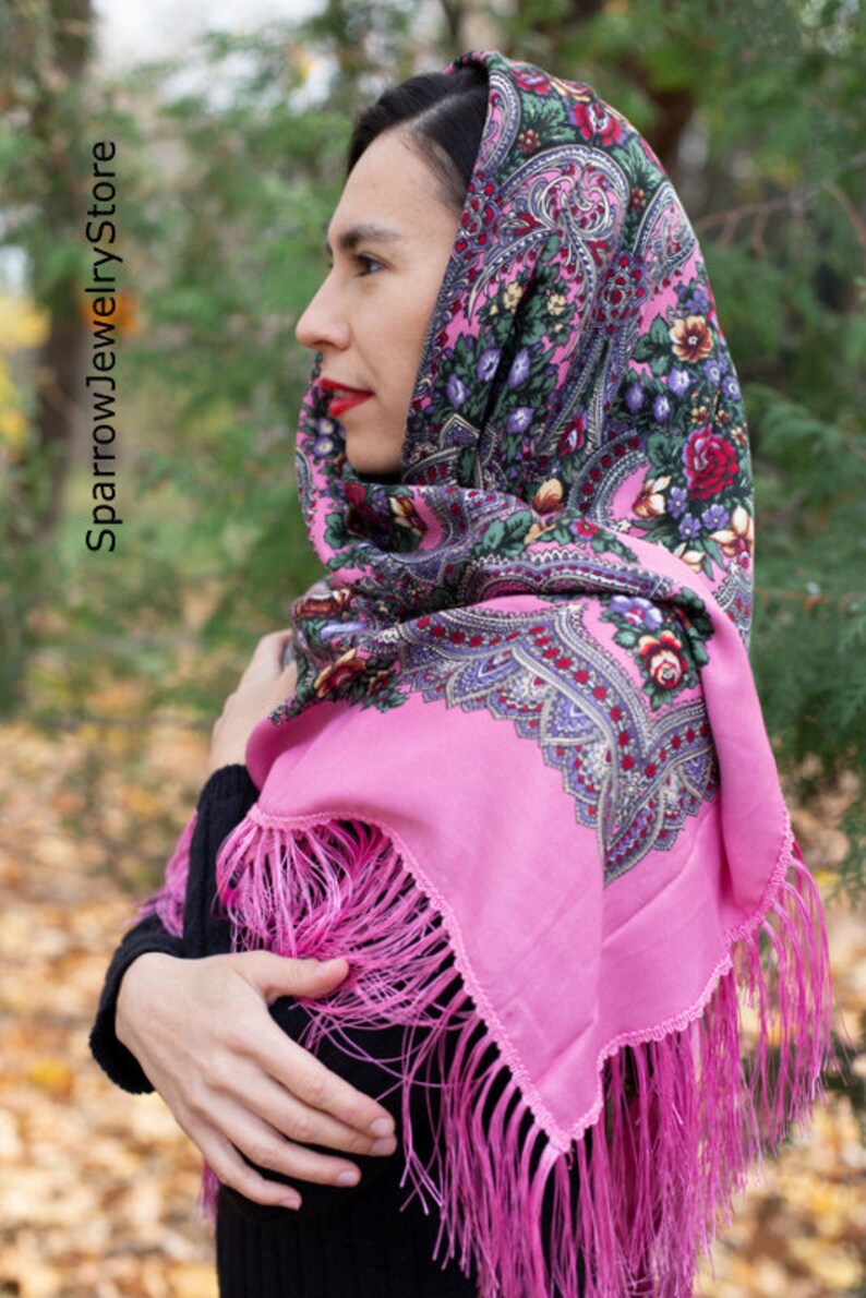May include: A pink headscarf with a floral pattern and fringe. The scarf is worn by a person with dark hair.