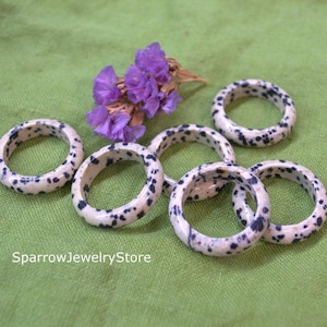 Puede incluir: Seis anillos de piedra natural moteados en blanco y negro. Los anillos están apilados sobre una superficie verde.