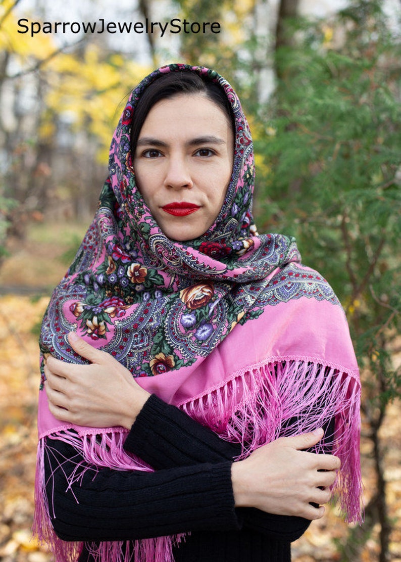 May include: A pink headscarf with a floral pattern and fringe. The scarf is worn by a person with dark hair and red lipstick.
