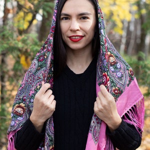 May include: A pink and floral patterned headscarf with fringe. The scarf is worn over the head and shoulders of a person with long dark hair. The person is wearing a black sweater.