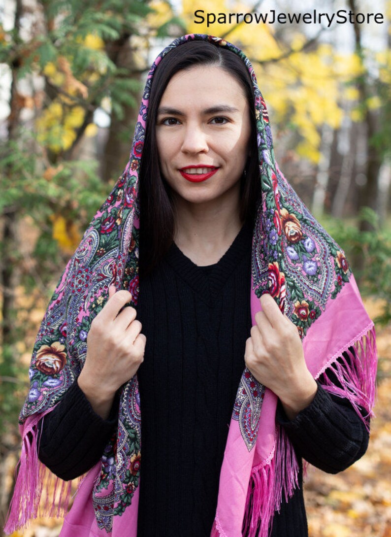 May include: A pink and floral patterned headscarf with fringe. The scarf is worn over the head and shoulders of a person with long dark hair. The person is wearing a black sweater.