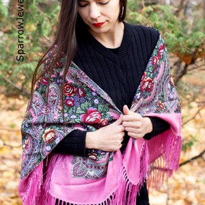 May include: A woman wearing a pink and black patterned shawl with floral designs and fringe. The shawl is draped over her shoulders and tied at the front.