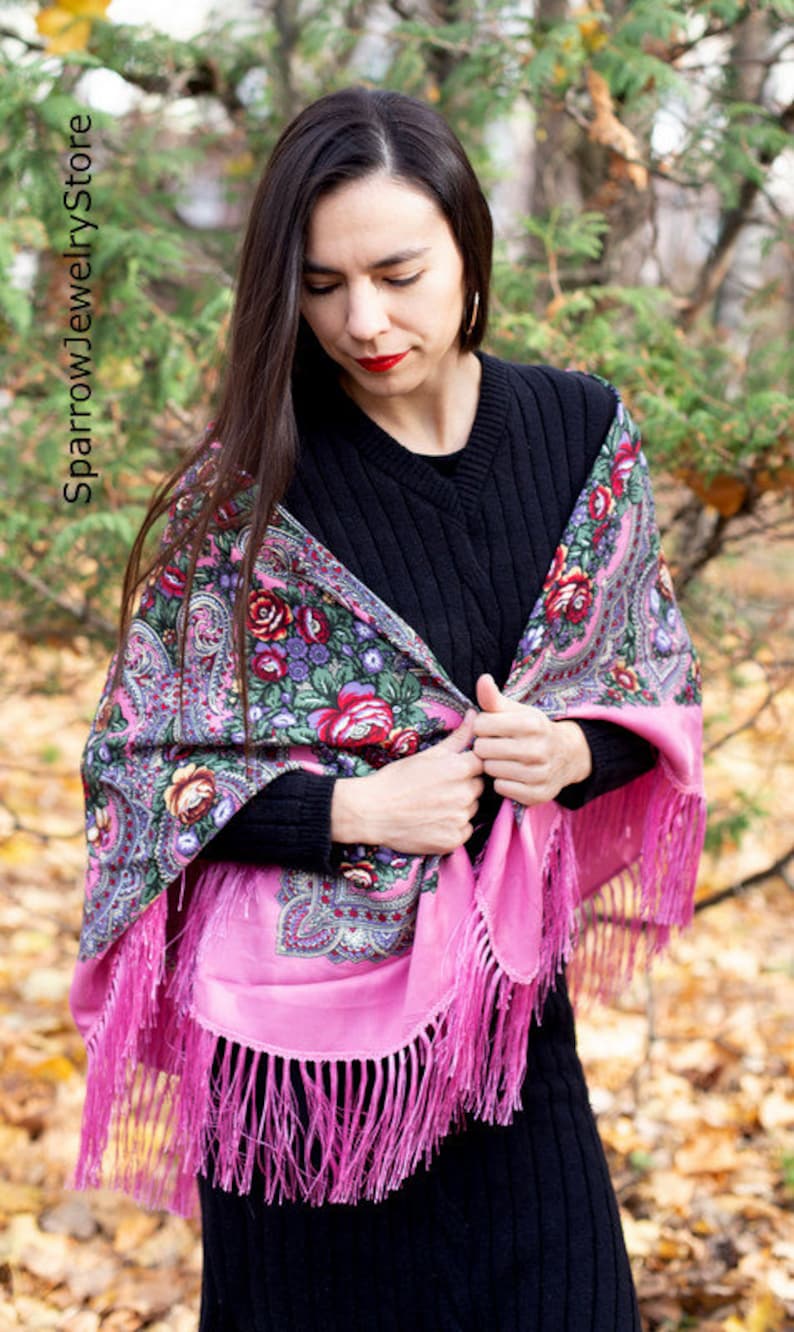 May include: A woman wearing a pink and black patterned shawl with floral designs and fringe. The shawl is draped over her shoulders and tied at the front.