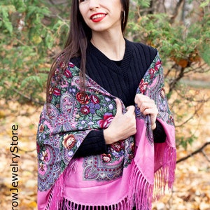 May include: A woman wearing a black sweater and a pink and floral patterned shawl with fringe. The shawl is draped over her shoulders and she is holding it in her hands. She is standing in front of a background of fall foliage.