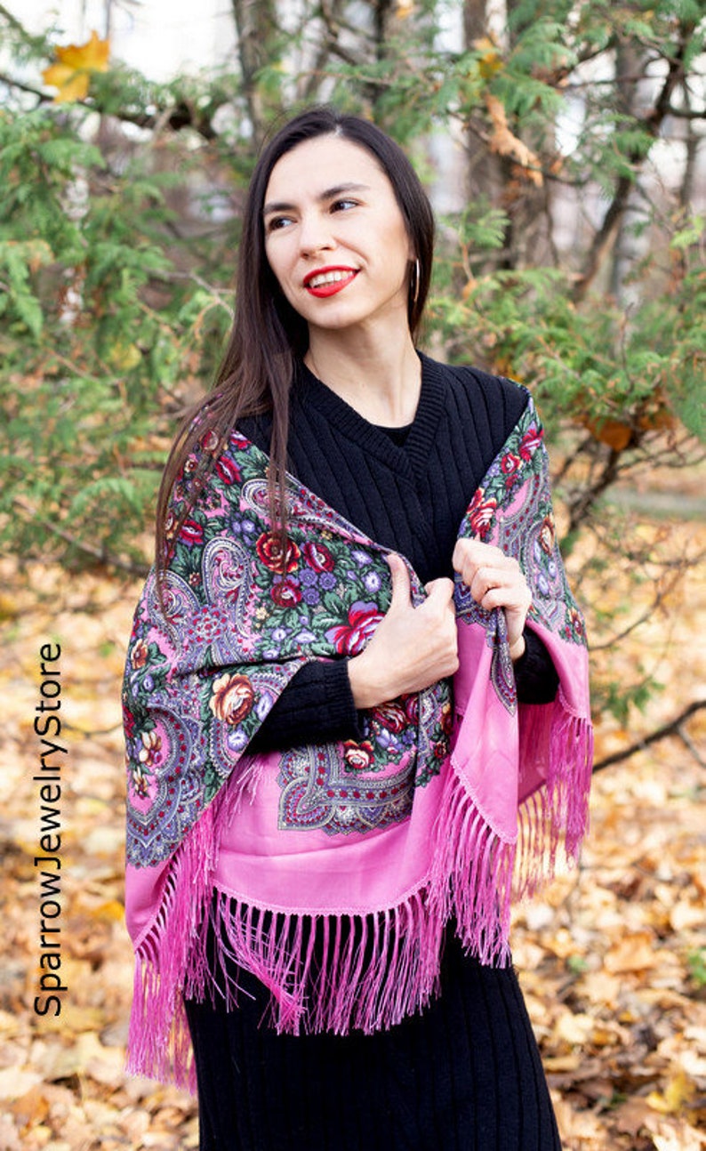 May include: A woman wearing a black sweater and a pink and floral patterned shawl with fringe. The shawl is draped over her shoulders and she is holding it in her hands. She is standing in front of a background of fall foliage.
