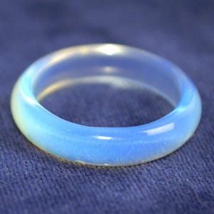 Könnte beinhalten: Ein durchscheinender, opalisierender Ring mit glatter, polierter Oberfläche. Der Ring wechselt von einem blassen Gelbton zu einem sanften Blau. Der Ring ist vor einem dunkelblauen, strukturierten Hintergrund platziert.