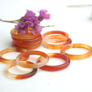 May include: A set of seven carnelian rings, stacked and arranged on a white surface. The rings are made of orange and red carnelian stone and have a polished finish.