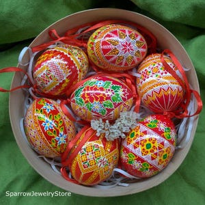 Puede incluir: Una caja redonda de madera llena de siete huevos de Pascua decorados. Los huevos presentan intrincados diseños en rojo, amarillo, blanco y verde, con cintas rojas. Se encuentran sobre papel blanco, sobre un fondo verde.