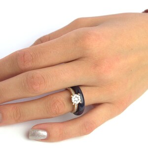 May include: A close-up of a hand wearing a ring with a large, round, clear stone surrounded by smaller stones. The ring is set in a gold band and is worn on the ring finger. The hand is also wearing a dark blue ring on the same finger.
