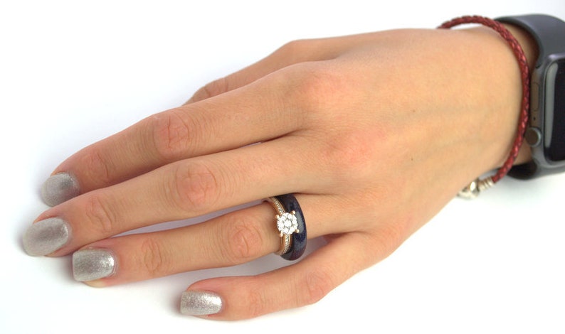 May include: A close-up of a hand wearing a ring with a large, round, clear stone surrounded by smaller stones. The ring is set in a gold band and is worn on the ring finger. The hand is also wearing a dark blue ring on the same finger.