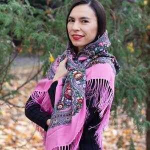 May include: A woman wearing a pink shawl with floral embroidery and fringe. The shawl is tied around her head and neck. She is standing in front of a green bush with fallen leaves on the ground.
