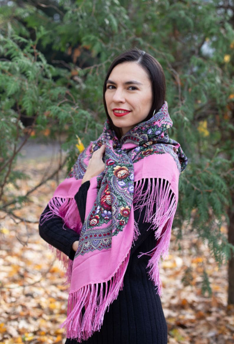 May include: A woman wearing a pink shawl with floral embroidery and fringe. The shawl is tied around her head and neck. She is standing in front of a green bush with fallen leaves on the ground.
