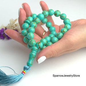 May include: A turquoise blue mala prayer bead necklace with a light blue tassel. The beads are round and have a marbled pattern. The tassel is made of soft fabric and has a rainbow colored thread.