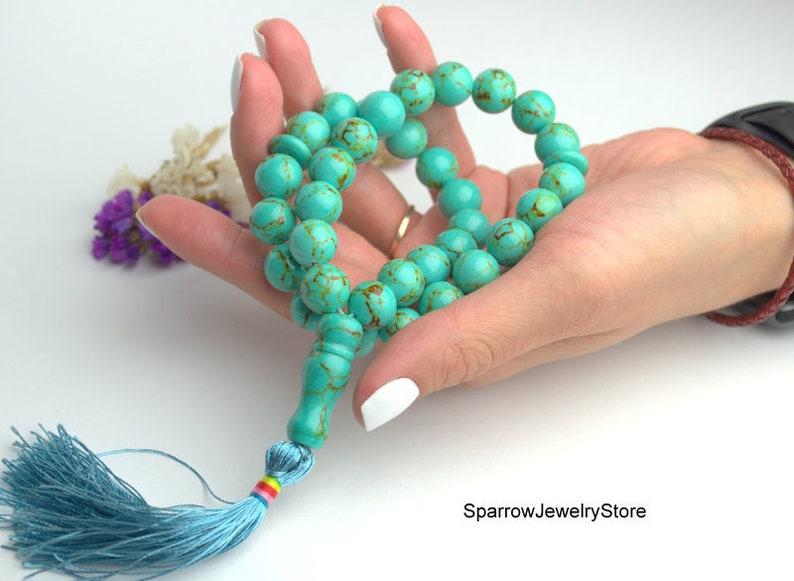 May include: A turquoise blue mala prayer bead necklace with a light blue tassel. The beads are round and have a marbled pattern. The tassel is made of soft fabric and has a rainbow colored thread.