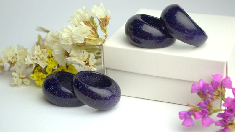 May include: Three dark blue rings with a sparkly texture, displayed on a white box with dried flowers on either side.