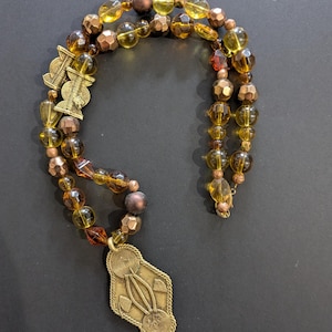 May include: A necklace featuring a series of amber-colored translucent beads, metallic bronze-toned faceted beads, and matte brown spherical beads. The piece includes two ornate, flat, brass-colored metal charms with spiral and geometric engravings.