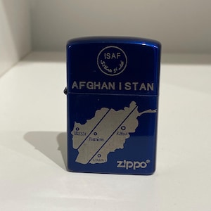 May include: A blue Zippo lighter with the word "AFGHANISTAN" and a map of Afghanistan. The lighter also features the text "ISAF" in a circle and the Zippo logo.