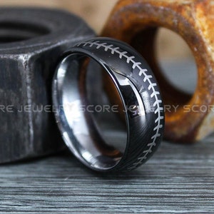 May include: A black tungsten carbide ring with a baseball stitch design. The ring is on a wooden surface with a rusty nut.