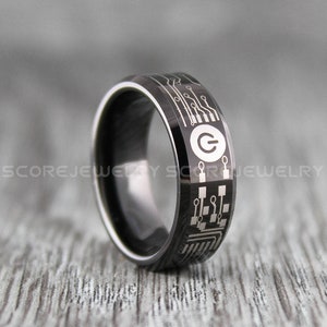 May include: A black tungsten carbide ring with a circuit board design and a power button symbol. The ring is on a wooden surface.