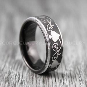May include: A black ring with a silver spade and floral pattern. The ring has a silver edge and is set against a grey background. The ring is a piece of jewellery.