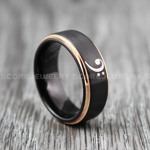Peut inclure: Bague en carbure de tungstène noir avec une bande en or rose et un symbole de clé de fa en blanc.