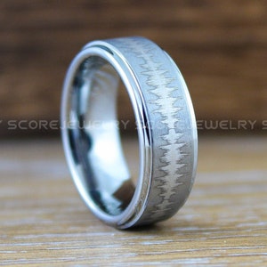 Puede incluir: Un anillo de carburo de tungsteno plateado con acabado cepillado y borde acanalado. El anillo tiene un sutil patrón de líneas grabadas en la superficie.