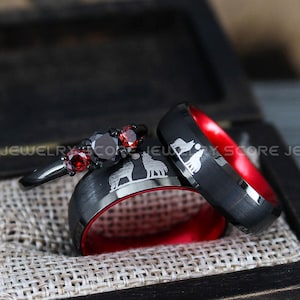 May include: A black ring box with red velvet lining, containing two black tungsten carbide rings with red inner bands. The rings feature an engraved design of a wolf howling at the moon. A smaller ring with three red gemstones is also visible in the box.