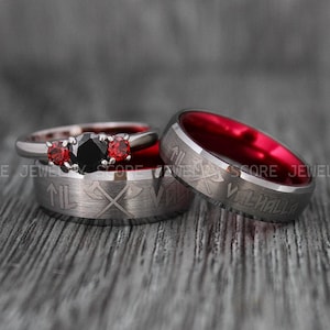 May include: A set of two wedding bands in black and red. The woman's band features a black gemstone with two smaller red gemstones on either side. The man's band is a tungsten carbide ring with a red interior and the inscription "Valhalla" in a silver font. Both bands feature a design of two crossed axes.