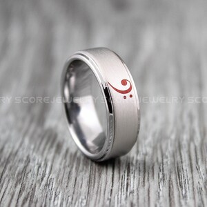 Peut inclure: Une bague en carbure de tungstène argentée avec une finition brossée et un symbole de clé de fa basse rouge gravé sur la bande.