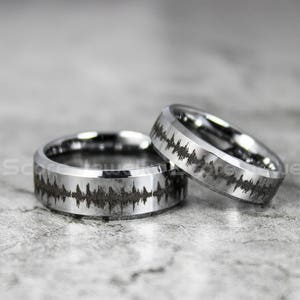 Op de afbeelding: Twee zilveren ringen van wolframcarbide met een zwart geluidgolfontwerp. De ringen zijn afgeschuind en hebben een gepolijste afwerking.