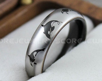 May include: A silver ring with black orca whale designs. The ring is displayed in a wooden box with a white interior. The ring has a polished finish and a dark interior. The orca whales are etched into the band.