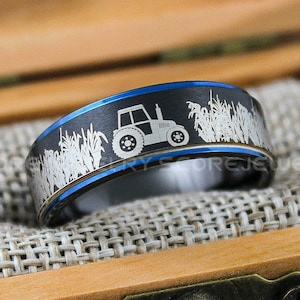 May include: A black and blue tungsten carbide ring with a white engraved design of a tractor in a field of corn.