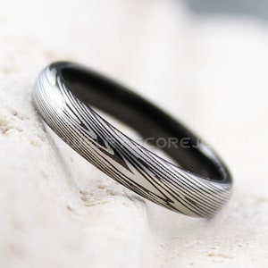 Pode incluir: Uma aliança de casamento com padrão prateado e preto. A aliança é feita de metal e tem um acabamento escovado.
