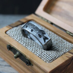 May include: A silver tungsten carbide ring with a black and white engraved image of a fish. The ring is in a wooden box with a brown fabric lining.