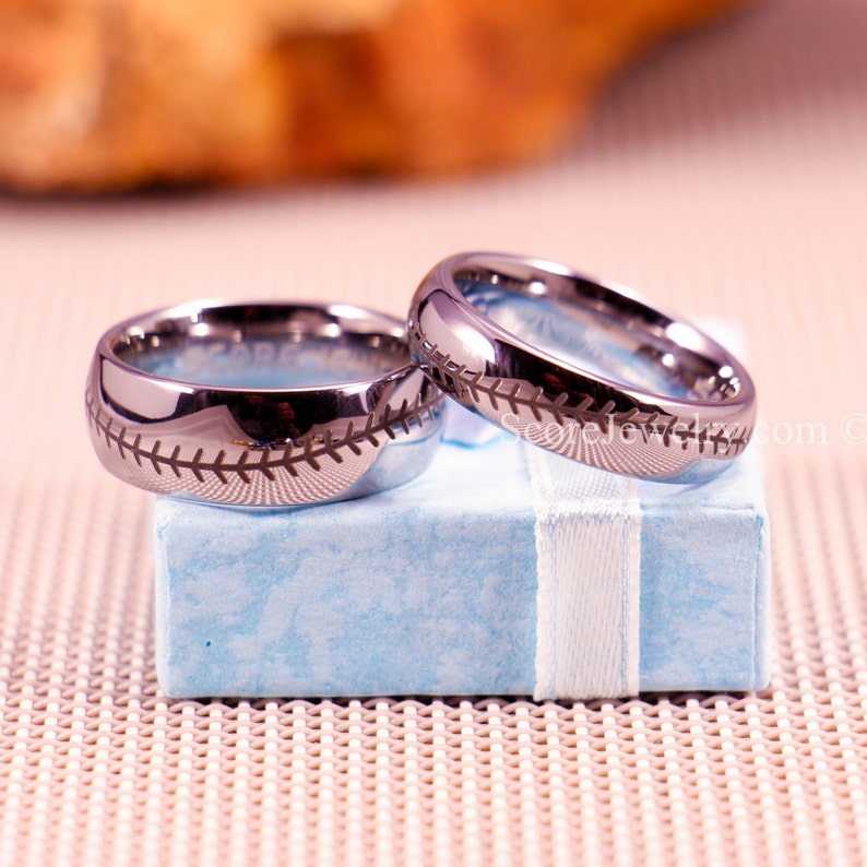 K&ouml;nnte beinhalten: Zwei silberne Eheringe mit einem Baseball-Nahtdesign. Die Ringe liegen auf einer hellblauen Schachtel mit einem wei&szlig;en Band.