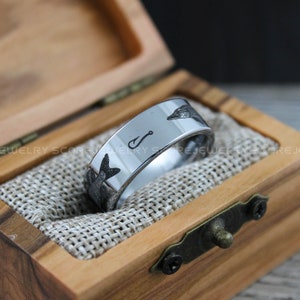 May include: A silver tungsten carbide ring with a fish and fishing hook design engraved on the band. The ring is in a wooden box with a brown fabric lining.