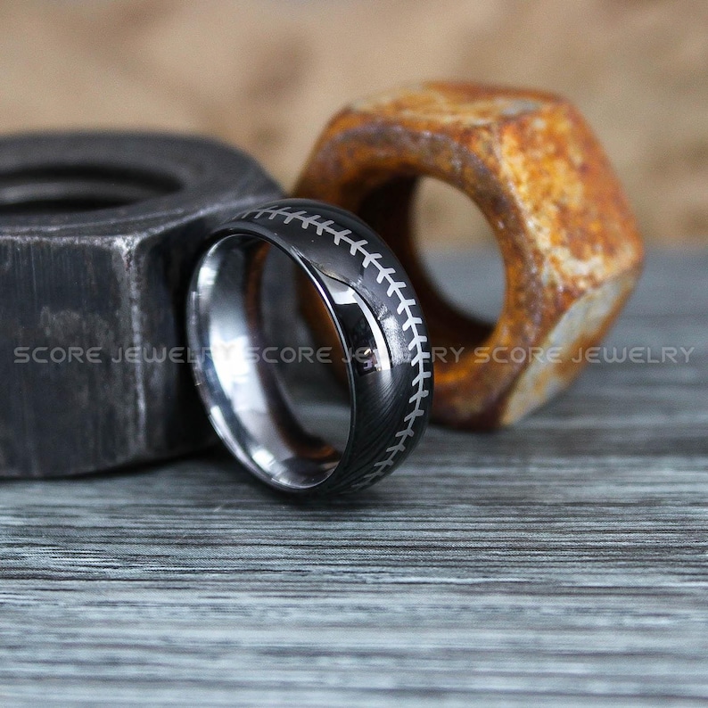 May include: A black tungsten carbide ring with a baseball stitch design. The ring is on a wooden surface with a rusty nut.
