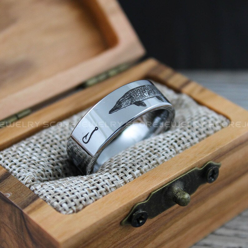 May include: A silver tungsten carbide ring with an engraved image of a fish and a fishing hook. The ring is in a wooden box.