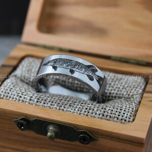 May include: A silver tungsten carbide ring with a detailed engraving of a fish on the band. The ring is displayed in a wooden box with a brown fabric lining.