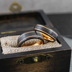 May include: Two black tungsten carbide wedding bands with gold inlay. The bands are in a wooden box with a brown fabric lining.