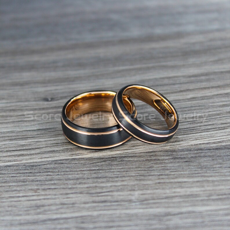 May include: Two black tungsten carbide wedding bands with a rose gold inner band. The bands are on a wood surface.
